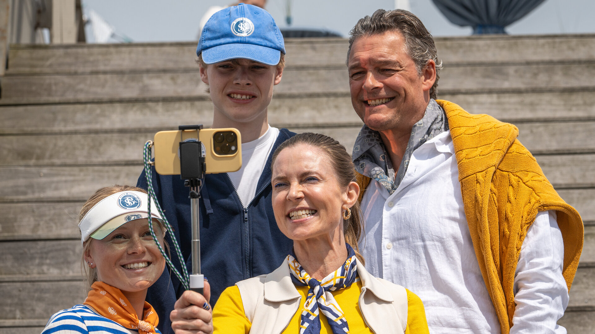 "Dan Sommerdahl - Tödliche Idylle: Tödlicher Ehrgeiz": Louise Busck (Camilla Gottlieb) nimmt mit ihren Kindern Frida (Rebekka Phillipson) und Karl (Mattias Brinch Nielsen) sowie ihrem Ehemann Jannik (Peter Reichhardt) auf einer Holztreppe im Hafen ein Foto auf. Louise hält ihr Smartphone in einem Selfiestick, alle lächeln in die Kamera.