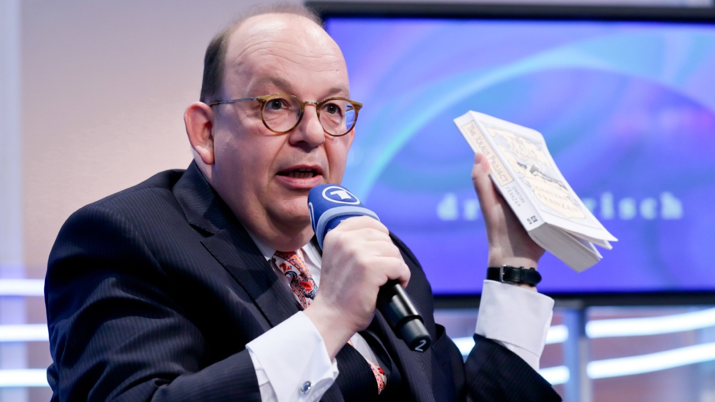 "Druckfrisch": Denis Scheck hält in seiner linken Hand ein Buch hoch und in der rechten Hand  ein Mikrofon vor seinen Mund. Hinter ihm steht ein Bildschrim.