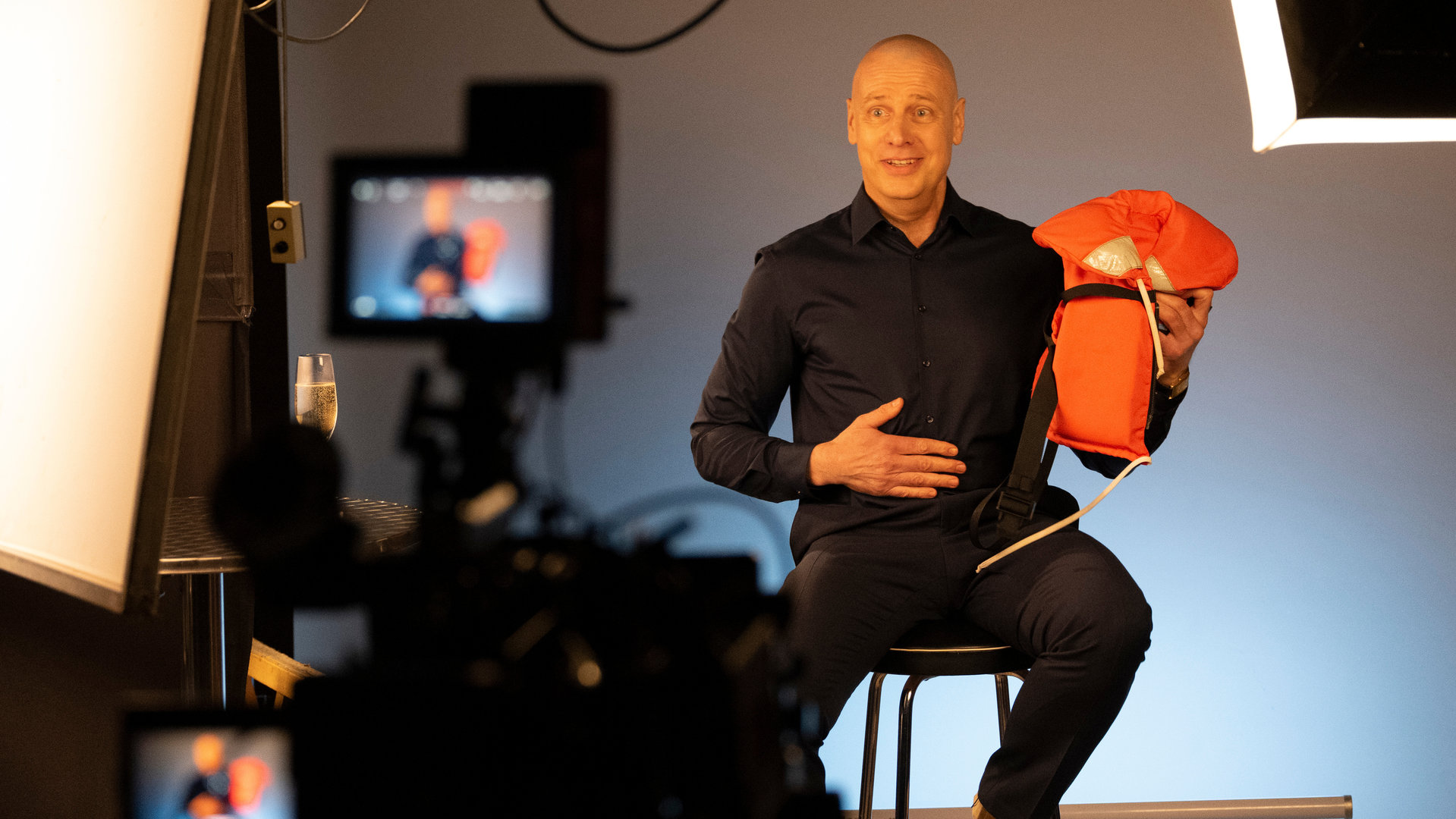 "SOKO Köln - Bis dass der Tod sie scheidet": Markus Hensch (Christoph Grunert) sitzt auf einem Hocker vor einer Kamera und hält einen orangenen Gegenstand in die Höhe. Er hält sich eine Hand an den Bauch und blickt angespannt lachend in die Kamera. Er trägt dunkle Kleidung. Die Wand hinter ihm ist grau und rechts im Bildrand steht ein Strahler, der auf ihn gerichtet ist. Vorne steht die aufgestellte Kamera.