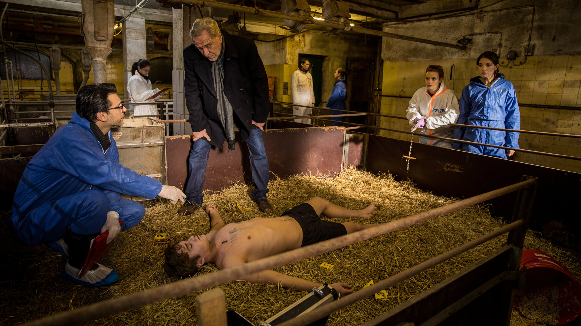 "SOKO München - Tod im Schweinestall": In einem Stall liegt eine Leiche auf dem Stroh, daneben steht Arthur Bauer (Gerd Silberbauer), am Kopf sitzt Dr. Weissenböck (Florian Odendahl) in der Hocke. Katharina Hahn (Bianca Hein) und Billie Curio (Sina Reiß) schauen vom Gelände zu. Im Hintergrund sind Komparsen.