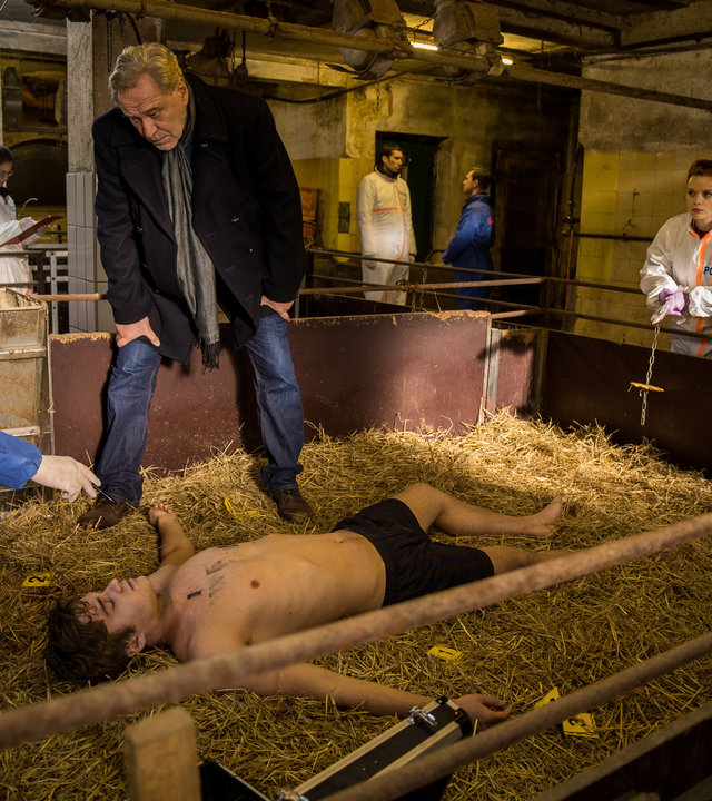 "SOKO München - Tod im Schweinestall": In einem Stall liegt eine Leiche auf dem Stroh, daneben steht Arthur Bauer (Gerd Silberbauer), am Kopf sitzt Dr. Weissenböck (Florian Odendahl) in der Hocke. Katharina Hahn (Bianca Hein) und Billie Curio (Sina Reiß) schauen vom Gelände zu. Im Hintergrund sind Komparsen.