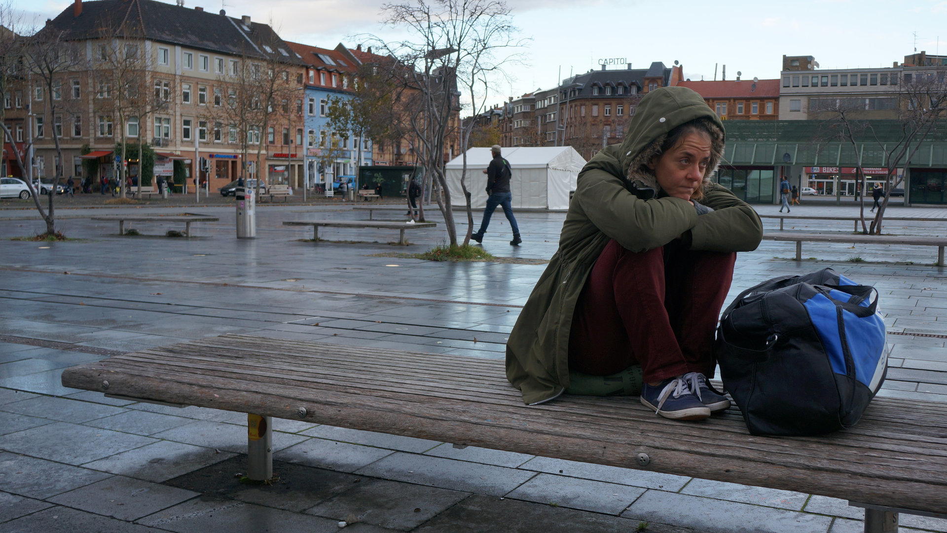 "Einfach Mensch - Auf der Straße": Bernadette L. sitzt auf einer Bank am Rande eines Marktplatzes.