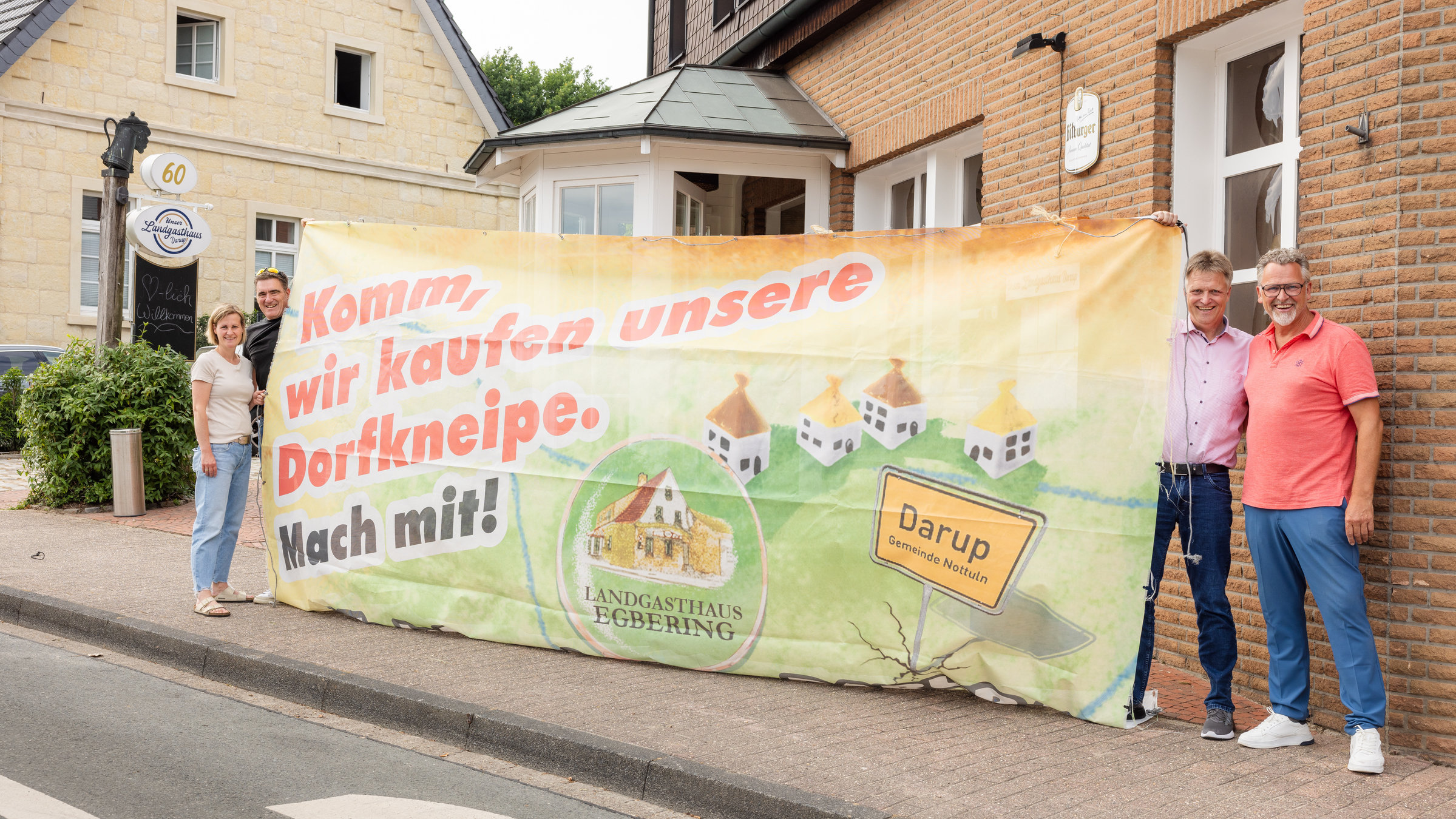 "37 °: Wir retten unser Dorf - Zusammenleben auf dem Land": Drei Männer und eine Frau stehen auf einem Bürgersteig. Je zwei links und zwei rechts im Bild. Dazwischen halten sie ein großes Banner mit der Aufschrift "Komm, wir kaufen unsere Dorfkneipe. Mach mit!" Im Hintergrund sieht man den Teil eines Hauses mit Klinkersteinen.