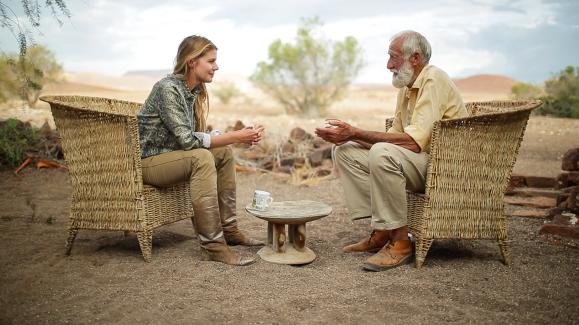 "Namib - Zauber der Wüste": Autorin Olivia Mokiejewski und Garth Owen Smith sitzen sich gegenüber in großen Korbsesseln, zwischen ihnen ein kleiner niedriger Holztisch, darauf steht eine Tasse. Sandboden, im Hintergrund (unscharf) braunes Gras, Steine, rötliche Dünen und vereinzelt grüne Büsche.