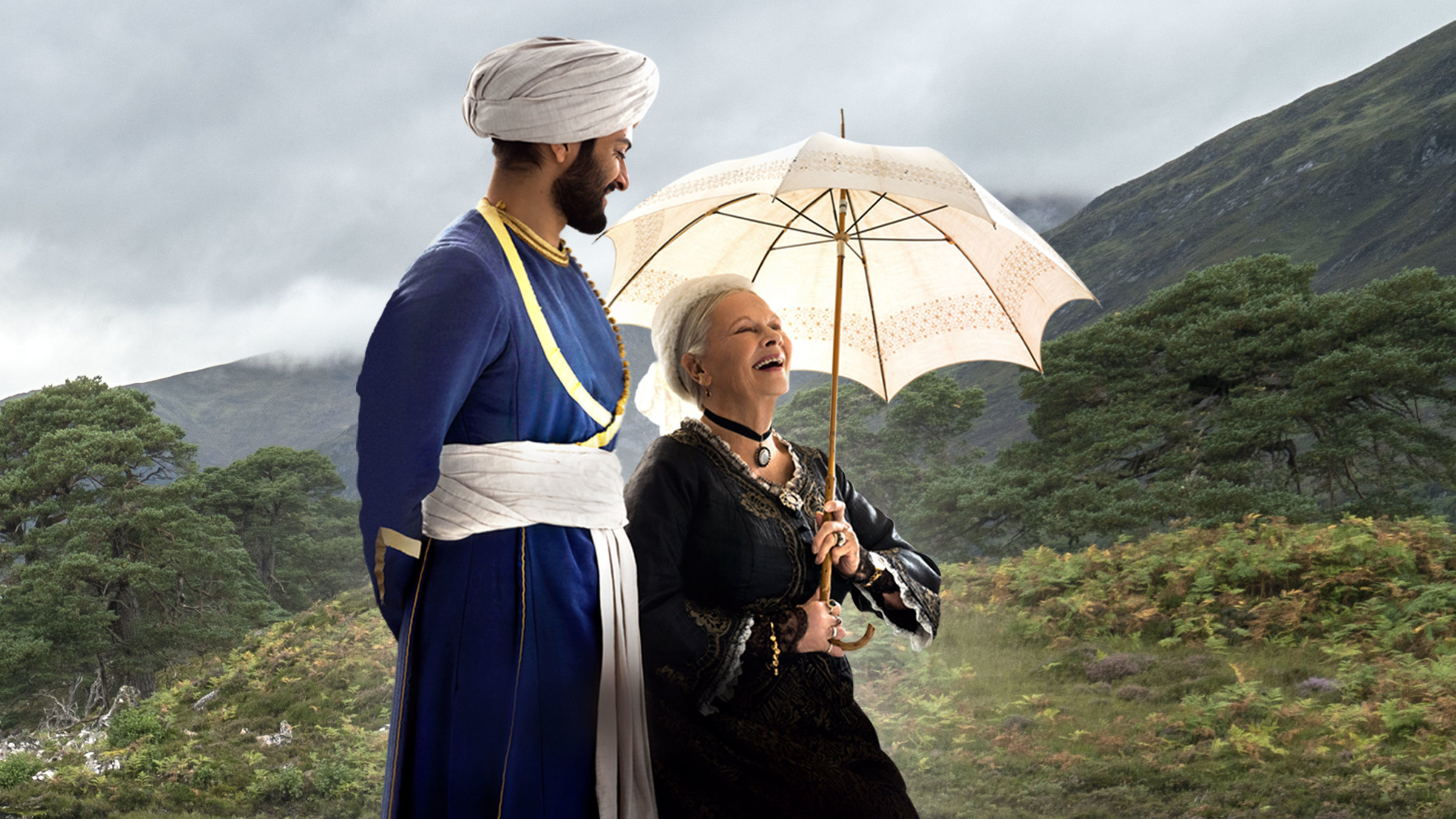 "Victoria und Abdul": Abdul Karim (Ali Fazal) und Queen Victoria (Judi Dench) spazieren nebeneinander über grün bewaldete Hügel. Abdul Karim schaut lächelnd zu Queen Victoria, die ihren aufgespannten Schirm über sich hält und herzhaft lacht.