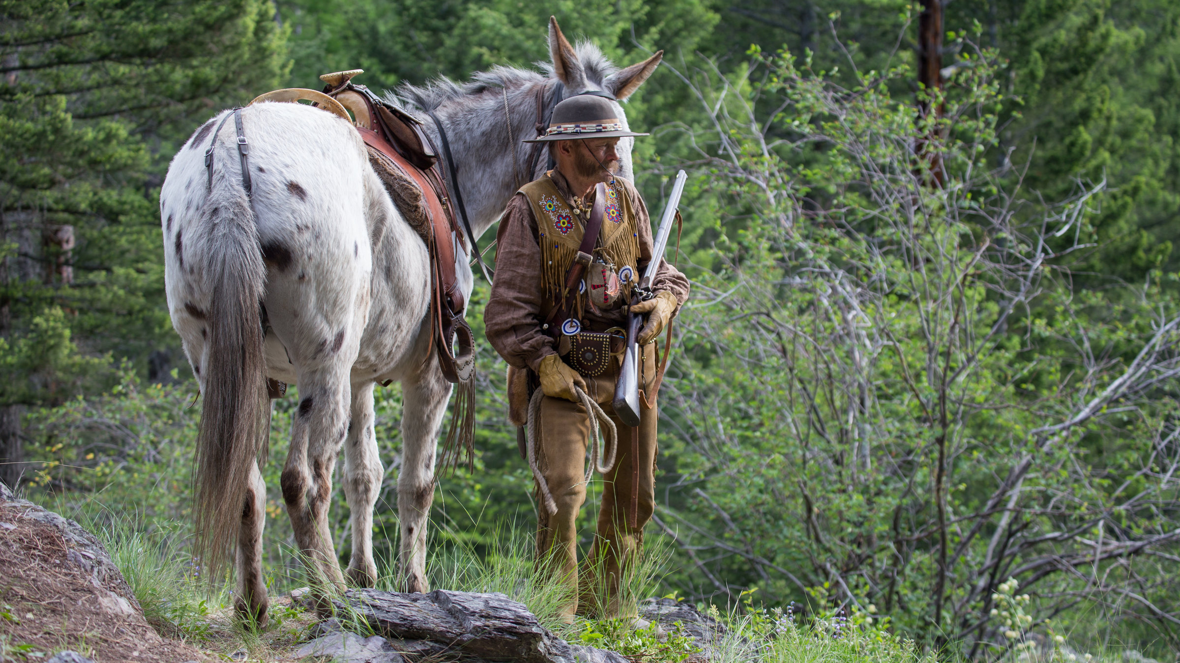 "Wilder Westen (1/3) - Gebirge": Lichtung im dichten Wald. In der Mitte des Bildes steht ein Trapper mit Gewehr in der Hand. Links von ihm ein Pferd, dessen Kopf hinter dem Trapper verschwindet. Rechts ein abgestorbener Busch.