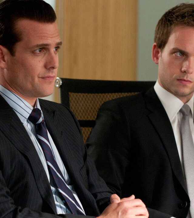 "Suits - Ausgehandelt": Harvey (Gabriel Macht) und Mike (Patrick J. Adams) sitzen nebeneinander an einem Konferenztisch. Beide tragen Anzug und blicken aufmerksam in dieselbe Richtung.