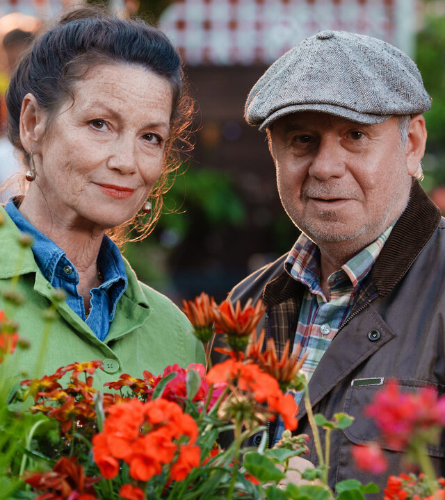 "Rosamunde Pilcher: Wer immer du bist": Im Hof der Gärtnerei: Inmitten von bunt blühenden Blumen stehen Charlotte Mansfield (Sabine Vitua) und Stephen Crayshaw (Joachim Król) nebeneinander und blicken lächelnd in die Kamera.