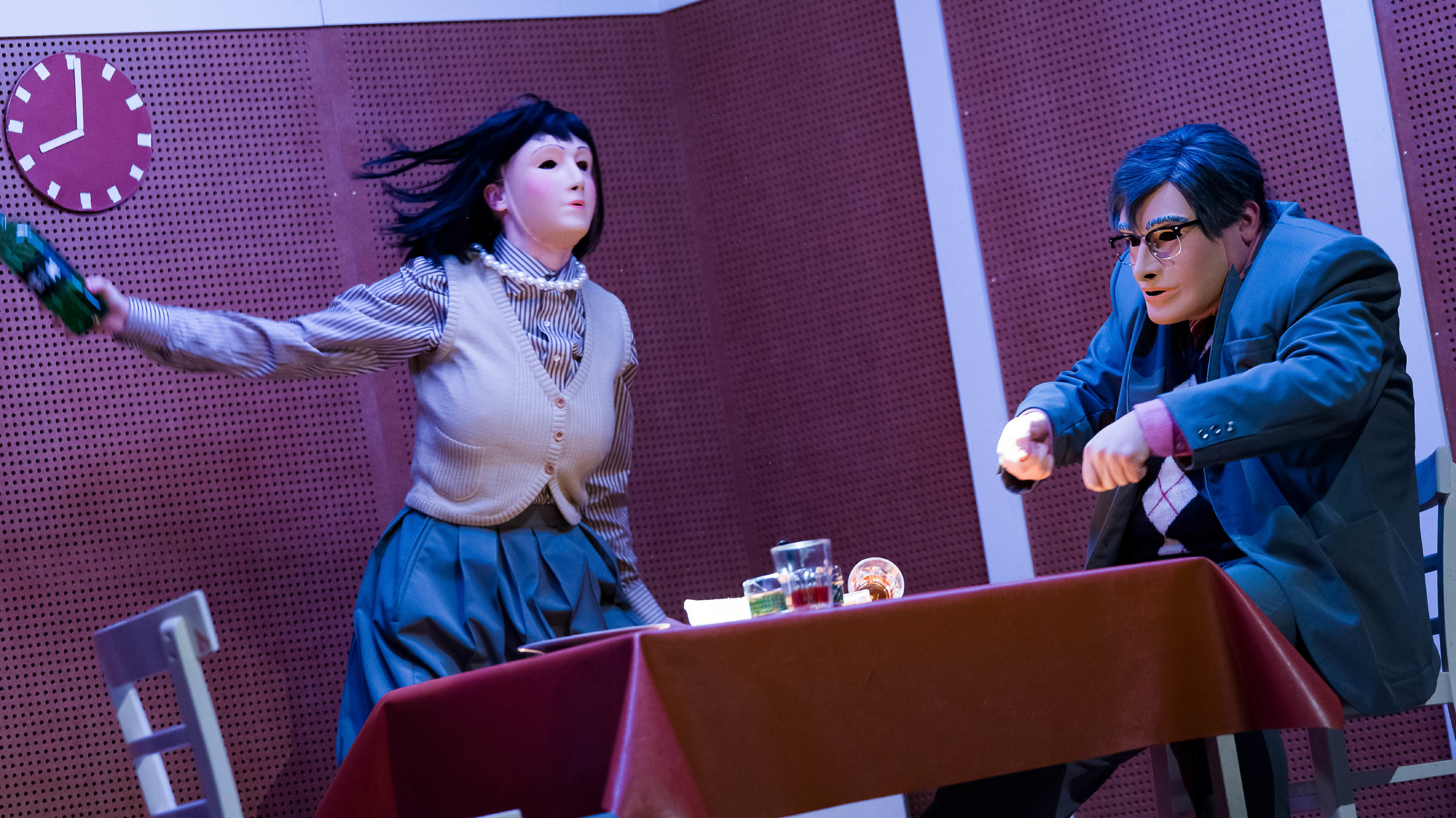 "Starke Stücke: humanistää! eine abschaffung der sparten": Im Vordergrund ein Tisch mit brauner Linoleumtischdecke, links ein leerer weißer Stuhl, rechts ein Mann sitzend, der die Hände über den Tisch hält als würde er essen schneiden. Er trägt einen grauen Anzug, eine dunkle Perücke und eine Brille. Sein Gesicht wird vollständig von einer Silikonmaske verdeckt. Hinter dem Tisch steht eine Frau, ebenfalls mit Maske, sie trägt eine schwarze Perücke, eine grau-weiß gestreifte Bluse, eine helle Strickweste, einen grauen Rock und eine dicke Perlenkette um den Hals. In der rechten Hand hält sie eine dunkelgrüne Weinflasche, als würde sie nach dem Mann werfen wollen. Die Uhr an der Wand im Hintergrund zeigt acht Uhr an.