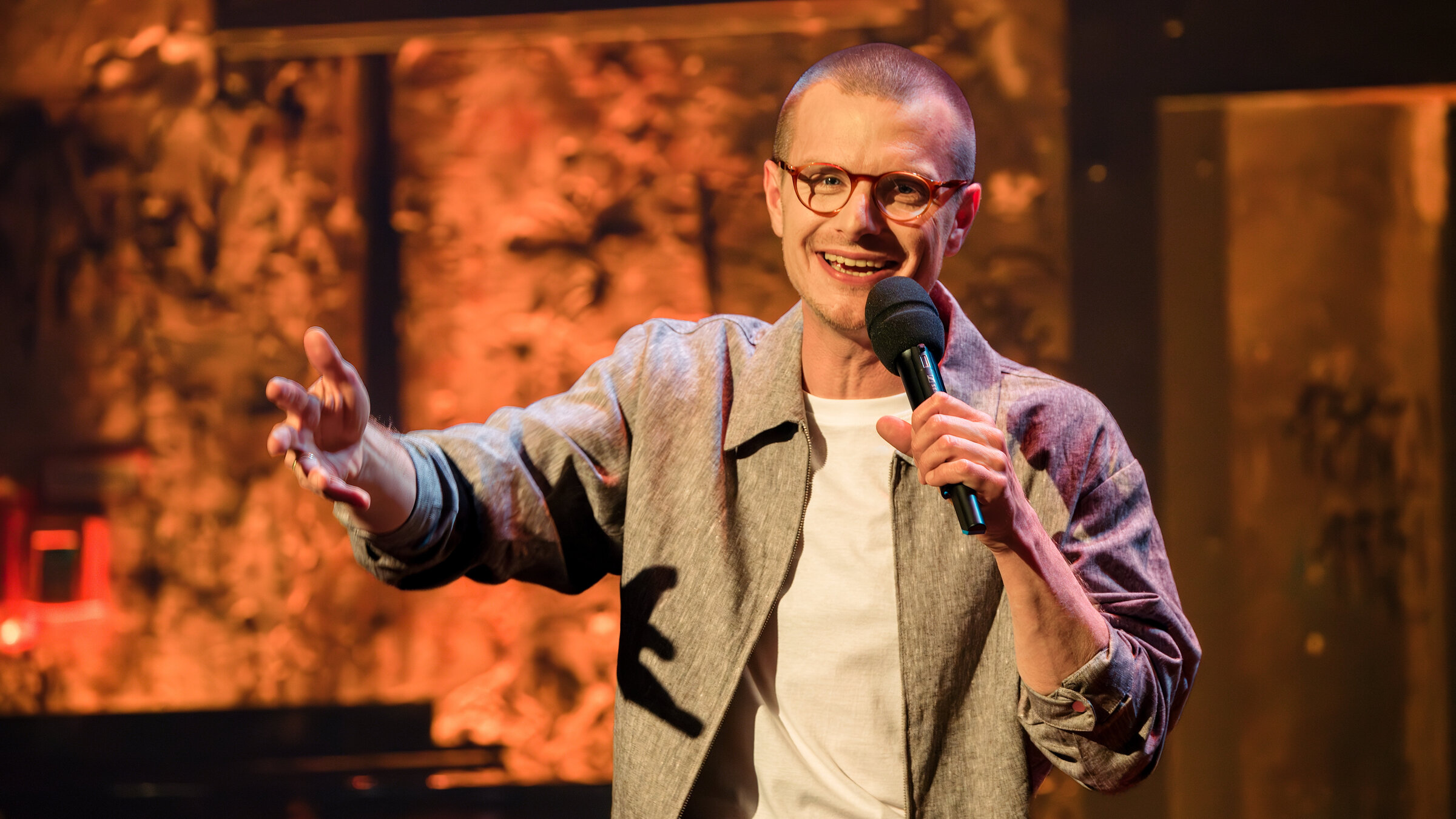 "Till Reiners` Happy Hour - Takeover - Primetime-Comedy mit Moritz Neumeier und Gästen": Der Stand-Up-Comedian und Gastmoderator Moritz Neumeier steht mit einem Mikrofon in der Hand auf der Bühne und lächelt Richtung Publikum.
