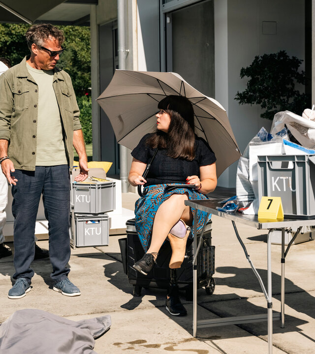 "SOKO Köln - Moneten erbeten": Dr. Pilar Westphal (Yulia Yáñez Schmidt) sitzt an einem Tatort auf einer Terrasse unter einem grauen Schirm auf einem schwarzen Rollkoffer. Sie blickt ernst zu Matti Wagner (Pierre Besson), der links von ihr steht und sie fragend ansieht. Rechts von Dr. Westphal steht eine Mitarbeiterin der Spurensicherung (Komparsin) in einem weißen Overall an einer geöffneten grauen Kiste mit der Aufschrift "KTU", die auf einem silbernen Metalltisch steht. Auf dem Terrassenboden sieht man ein Stück einer grauen Plane liegen. Hinter Matti steht ein weiterer Mitarbeiter der Spurensicherung (Komparse) in einem weißen Schutzanzug an einem geöffneten Koffer.