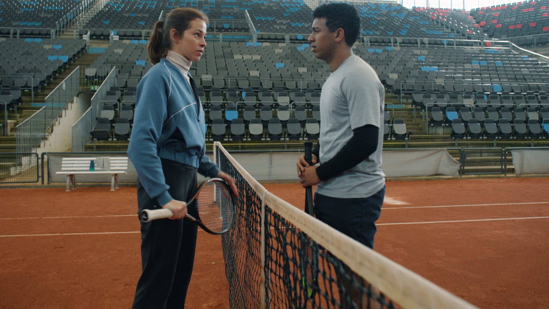 "Notruf Hafenkante - Dreamteam": Marie (Marie Hacke) und Ben (Aaron Maldonado-Morales) stehen sich auf einem Tennisplatz gegenüber. Das Netz des Platzes ist zwischen ihnen gespannt. Sie sehen sich an. Beide halten einen Tennisschläger in den Händen. Im Hintergrund liegt eine leere Tribüne