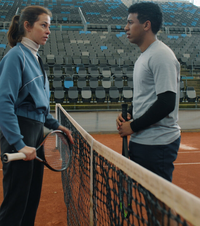 "Notruf Hafenkante - Dreamteam": Marie (Marie Hacke) und Ben (Aaron Maldonado-Morales) stehen sich auf einem Tennisplatz gegenüber. Das Netz des Platzes ist zwischen ihnen gespannt. Sie sehen sich an. Beide halten einen Tennisschläger in den Händen. Im Hintergrund liegt eine leere Tribüne