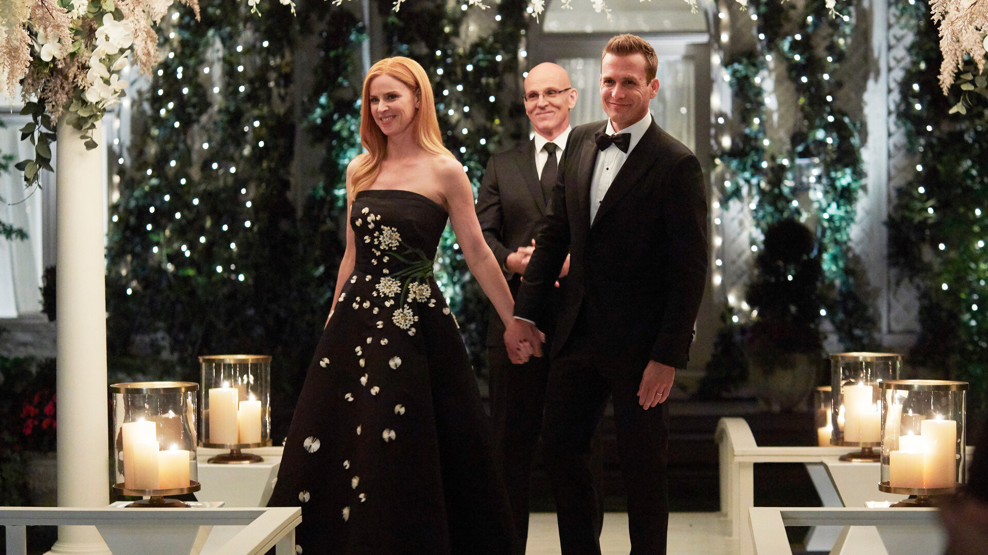 "Suits - Ein letzter Trick": Donna (Sarah Rafferty), in einem schwarzen mit Pailletten bestickten Kleid, und Harvey (Gabriel Macht) strahlen beide glücklich und halten sich an den Händen. Im Hintergrund steht ein Mann (Komparse) in einer traumhaften Blumen- und Lichterdekoration. Vor ihnen stehen auf einem Geländer vier Windlichter mit brennenden Kerzen.