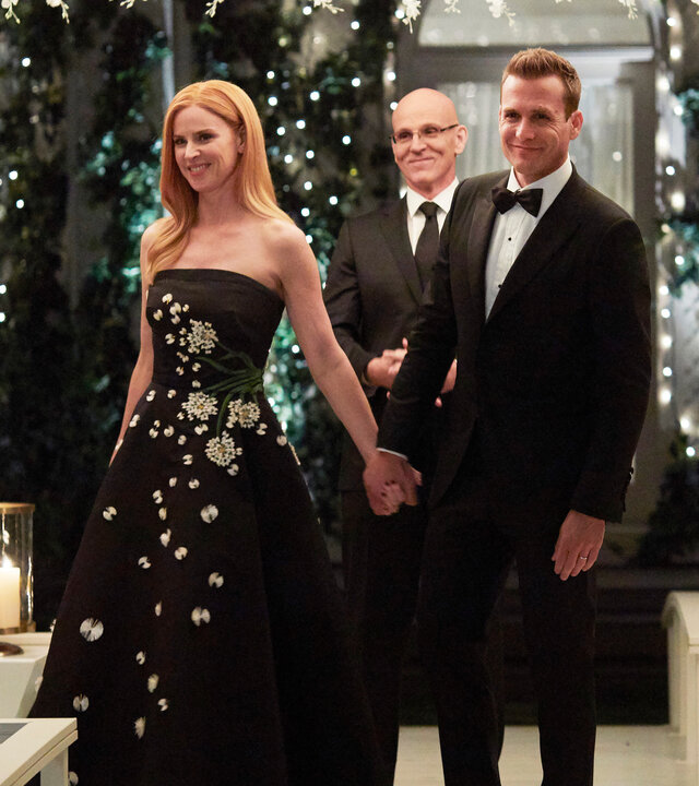 "Suits - Ein letzter Trick": Donna (Sarah Rafferty), in einem schwarzen mit Pailletten bestickten Kleid, und Harvey (Gabriel Macht) strahlen beide glücklich und halten sich an den Händen. Im Hintergrund steht ein Mann (Komparse) in einer traumhaften Blumen- und Lichterdekoration. Vor ihnen stehen auf einem Geländer vier Windlichter mit brennenden Kerzen.