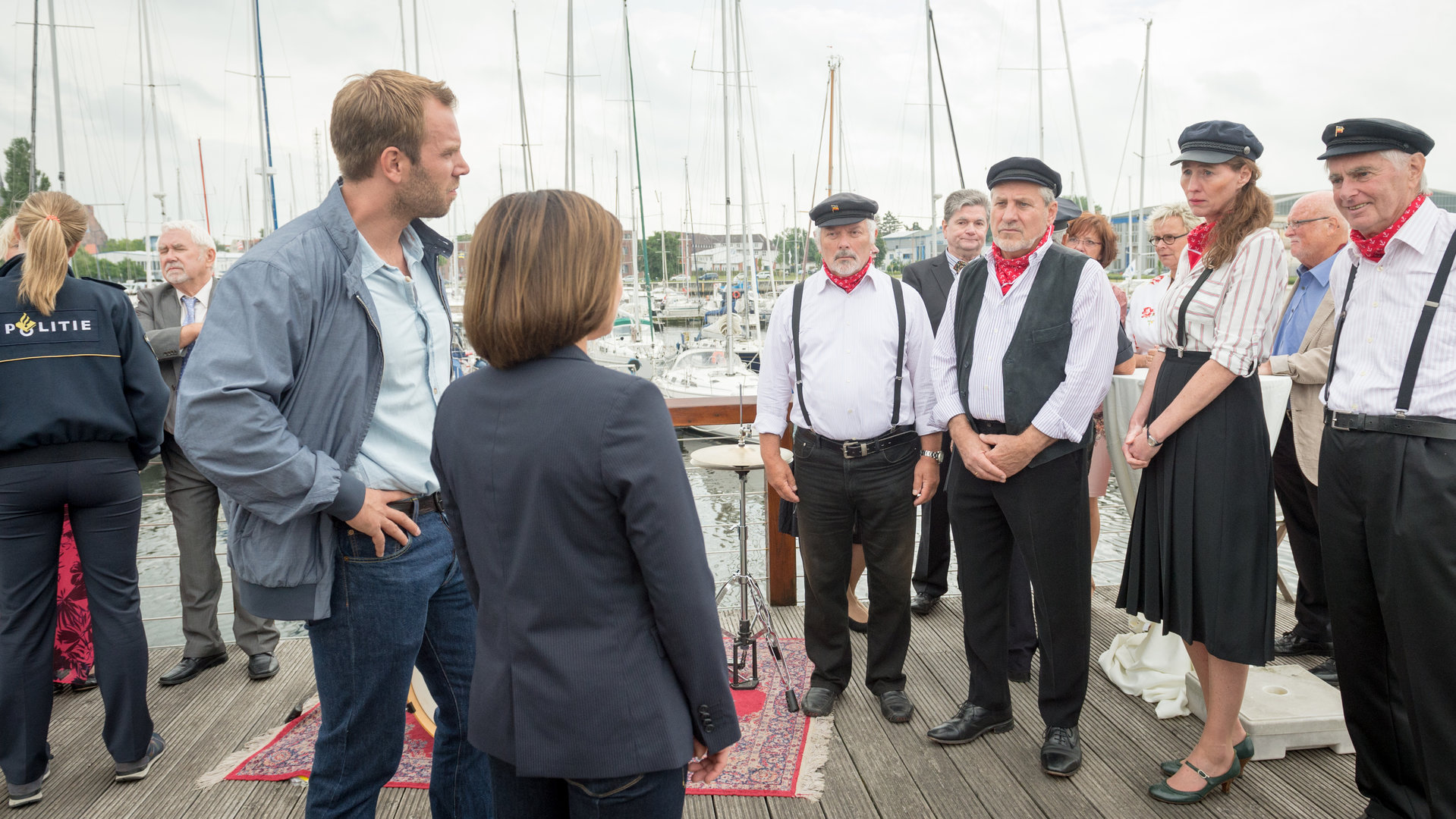 "SOKO Wismar – Eheversprechen": Lars Pöhlmann (Dominic Boeer) und Katrin Börensen (Claudia Schmutzler) stehen an einem Hafen vor einer Gruppe Männer und Frauen in Seemannstrachten. Im Hintergrund erkennt man festlich gekleidete Gäste (Komparsen). Auch Anneke van der Meer (Isabel Berghout) befragt die Anwesenden.