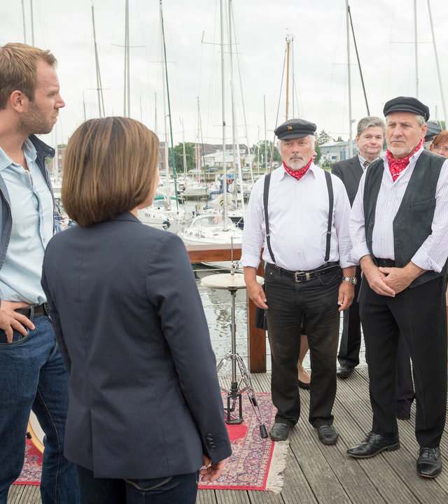 "SOKO Wismar – Eheversprechen": Lars Pöhlmann (Dominic Boeer) und Katrin Börensen (Claudia Schmutzler) stehen an einem Hafen vor einer Gruppe Männer und Frauen in Seemannstrachten. Im Hintergrund erkennt man festlich gekleidete Gäste (Komparsen). Auch Anneke van der Meer (Isabel Berghout) befragt die Anwesenden.