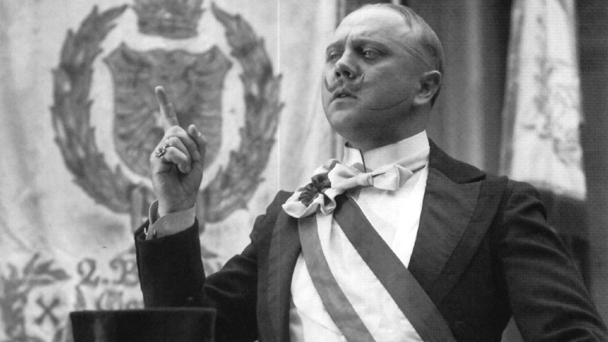 "Der Untertan": Diederich Heßling (Werner Peters) steht mit hochgezwirbeltem Schnurrbart hinter einem Rednerpult und reckt mit selbstgewissem Blick den Zeigerfinger. Er trägt ein festliches schwarzes Jackett und ein weißes Hemd mit Stehkragen, darüber eine dreifarbige Schärpe. Vor ihm auf dem Pult sind ein schwarzer Zylinder und weiße Handschuhe abgelegt. Im Hintergrund ist eine Fahne mit Kaiserwappen aufgehängt.