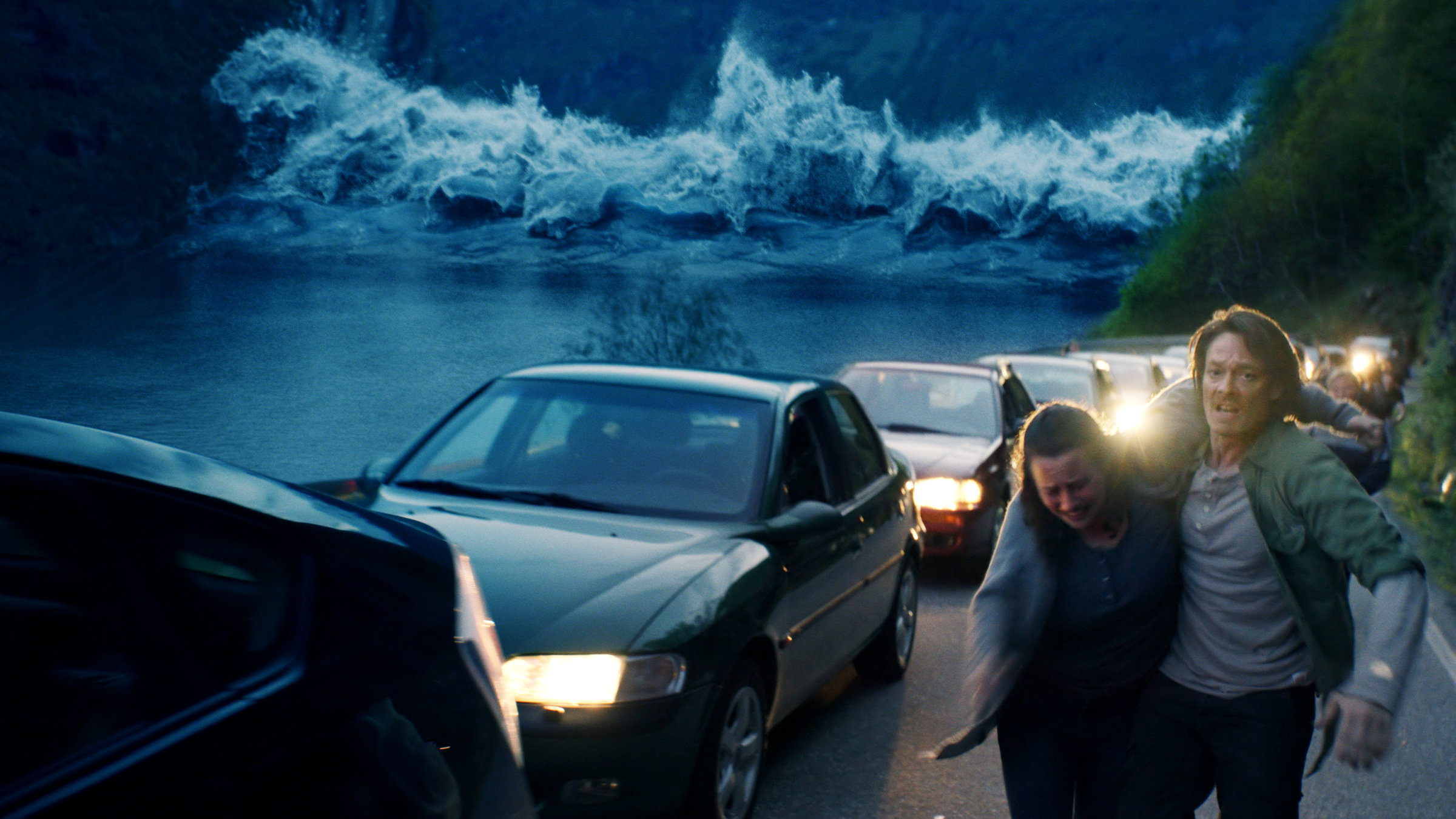 "The Wave - Die Todeswelle": Kristian (Kristoffer Joner) hält Anna (Silje Breivik) stützend im Arm. Sie befinden sich auf einer Straße, wobei sie vor der Flutwelle flüchten, die im Hintergrund sichtbar ist. Links neben ihnen stehen parkende Autos.
