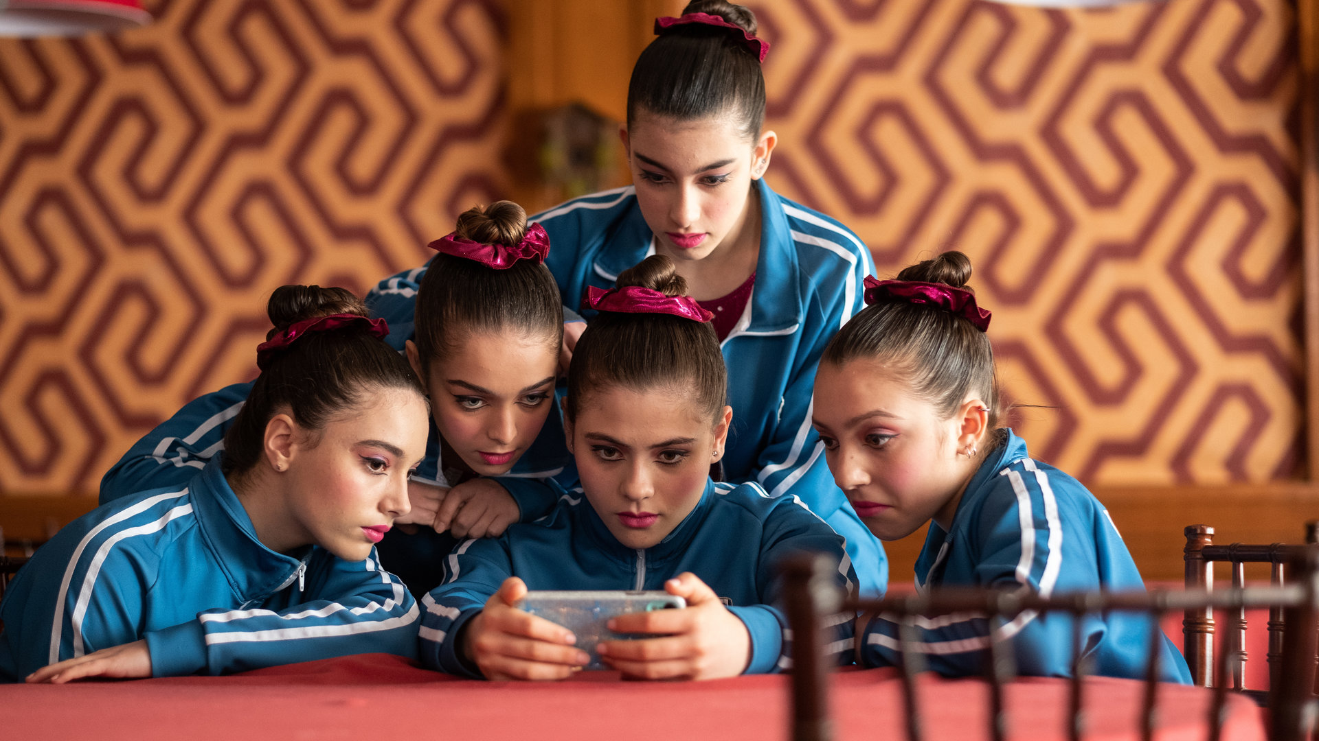 "The Gymnasts: Dienstag": Nadia (Federica Cuomo), Benedetta (Eva Iurlaro), Carla (Giada Savi), Martina (Alessia De Falco) und Anna (Giada Pirozzi) blicken gemeinsam auf ein Smartphone, das Carla in den Händen hält.