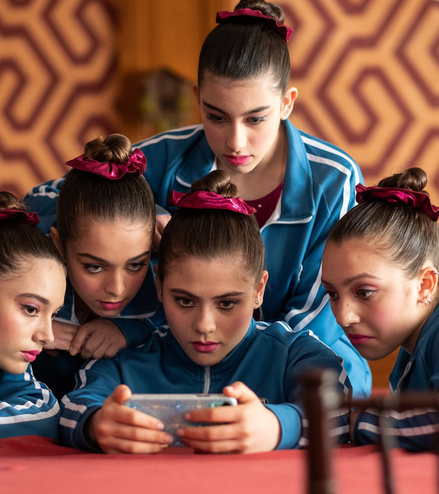 "The Gymnasts: Dienstag": Nadia (Federica Cuomo), Benedetta (Eva Iurlaro), Carla (Giada Savi), Martina (Alessia De Falco) und Anna (Giada Pirozzi) blicken gemeinsam auf ein Smartphone, das Carla in den Händen hält.