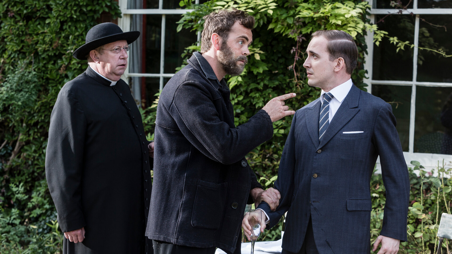 "Father Brown - Die Sünden der anderen": Father Brown (Mark Williams) steht hinter Sid Carter (Alex Price), der Giles Foster (David Reed) am Arm packt, mit dem Finger auf ihn zeigt und ihn drohend anschaut. Giles Foster hält ein Glas Champagner in der Hand, wendet sich leicht ab und schaut Sid Carter ängstlich ins Gesicht.