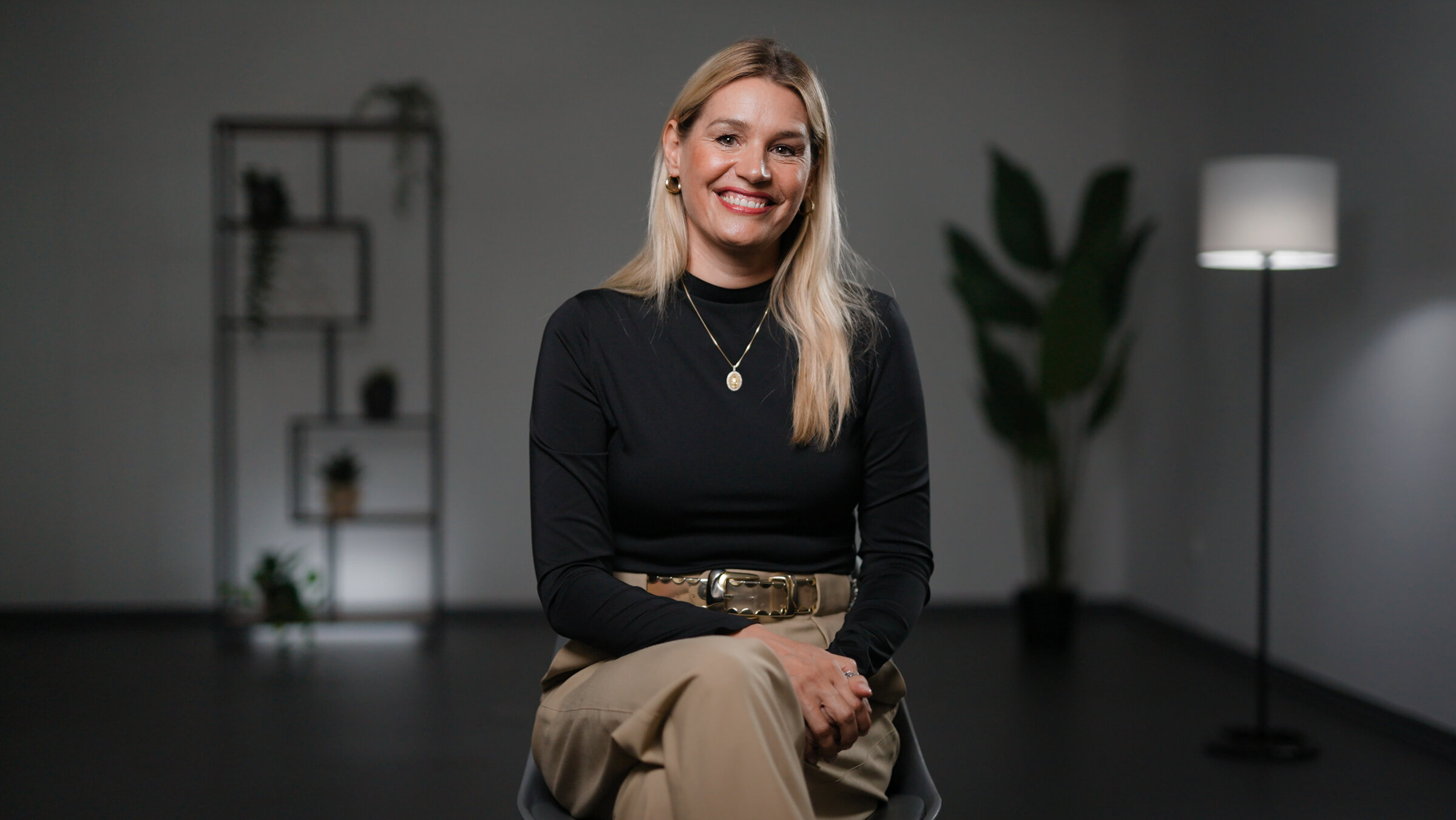 "True ART Crime – Spektakuläre Verbrechen in der Kunstwelt": Eine Frau sitzt auf einem Stuhl. Sie hat blonde Haare, trägt einen schwarzen Pullover und eine beige Hose. Die Beine verschränkt sie übereinander. Die Frau schaut direkt in die Kamera und lächelt. Im Hintergrund ist links ein beleuchtetes Regal zu sehen, rechts hinten eine Pflanze in der Ecke sowie eine Standleuchte.