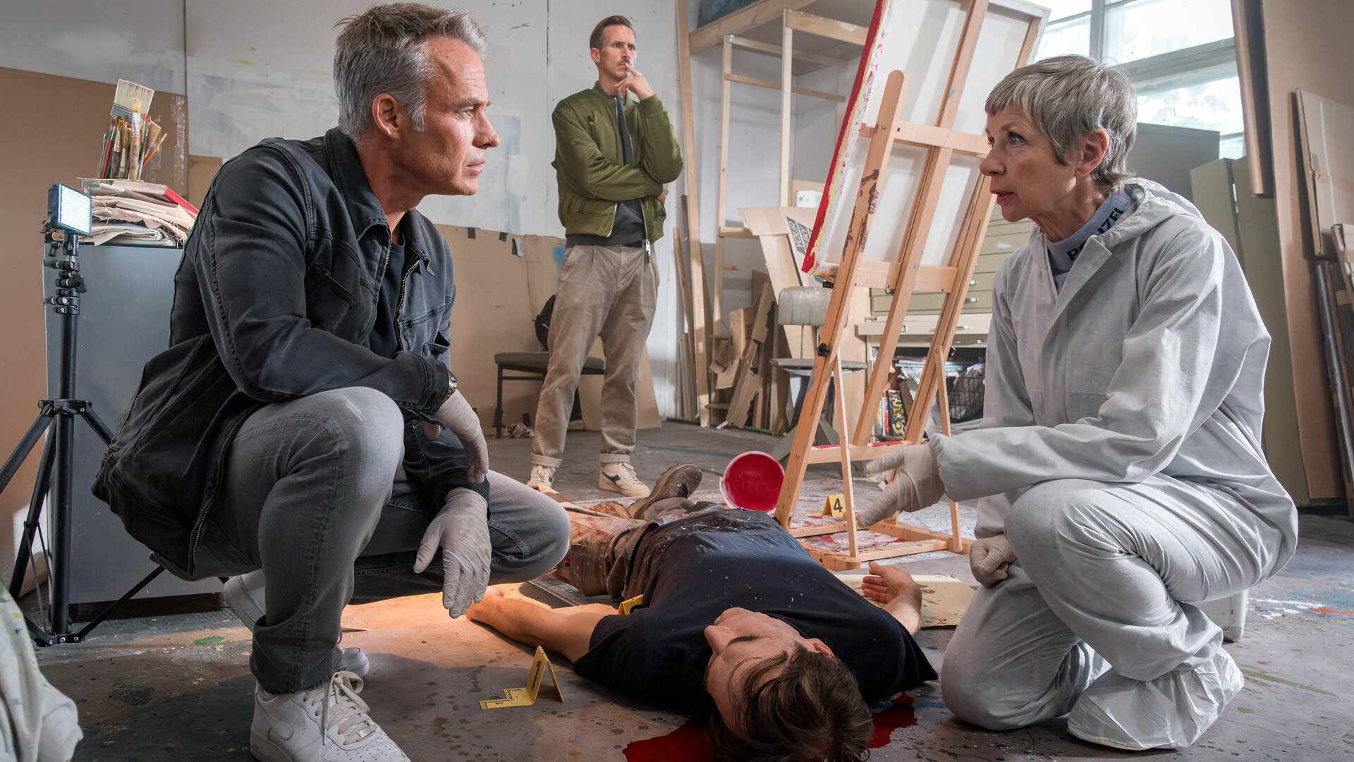 "SOKO Leipzig - Die Überwältigung": In einem großen, hellen Atelier: Jan Maybach (Marco Girnth) und Prof. Dr. Sabine Rossi (Anna Stieblich) hocken neben der Leiche von Maik Wuttke (Jonas Fürstenau), die am Boden vor einer Staffelei liegt. Währenddessen begutachtet Moritz Brenner (Johannes Hendrik Langer) im Hintergrund ein Bild auf einer Leinwand.