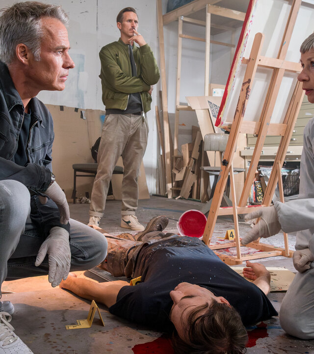 "SOKO Leipzig - Die Überwältigung": In einem großen, hellen Atelier: Jan Maybach (Marco Girnth) und Prof. Dr. Sabine Rossi (Anna Stieblich) hocken neben der Leiche von Maik Wuttke (Jonas Fürstenau), die am Boden vor einer Staffelei liegt. Währenddessen begutachtet Moritz Brenner (Johannes Hendrik Langer) im Hintergrund ein Bild auf einer Leinwand.