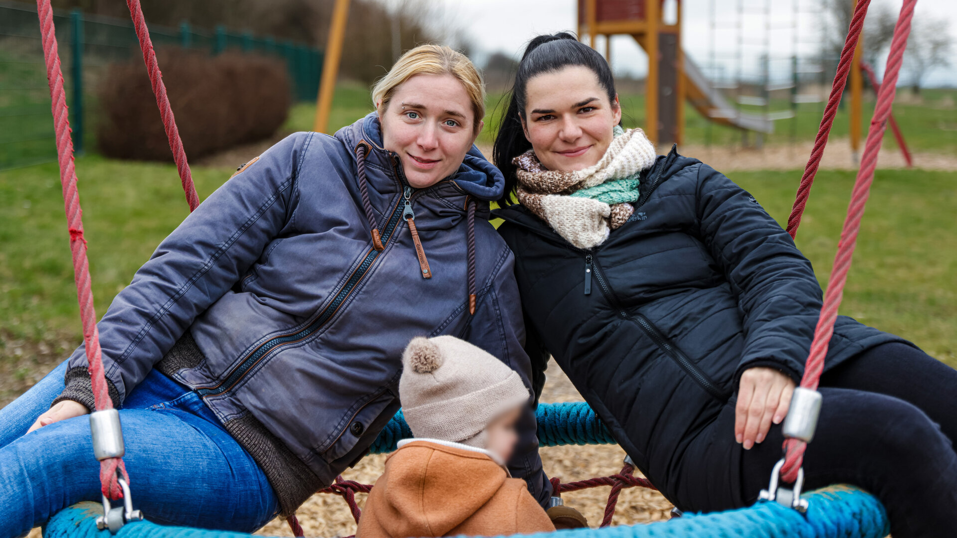 "TRU Stories: Gute Eltern - Zwei Mamas und ihr Kind": Lena und Patricia sitzen gemeinsam mit einem Kleinkind in einer Schaukel und lächeln in die Kamera. Im Hintergrund steht eine Rutsche.