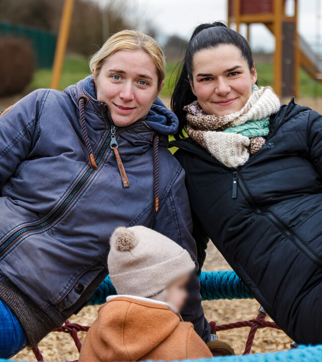 "TRU Stories: Gute Eltern - Zwei Mamas und ihr Kind": Lena und Patricia sitzen gemeinsam mit einem Kleinkind in einer Schaukel und lächeln in die Kamera. Im Hintergrund steht eine Rutsche.