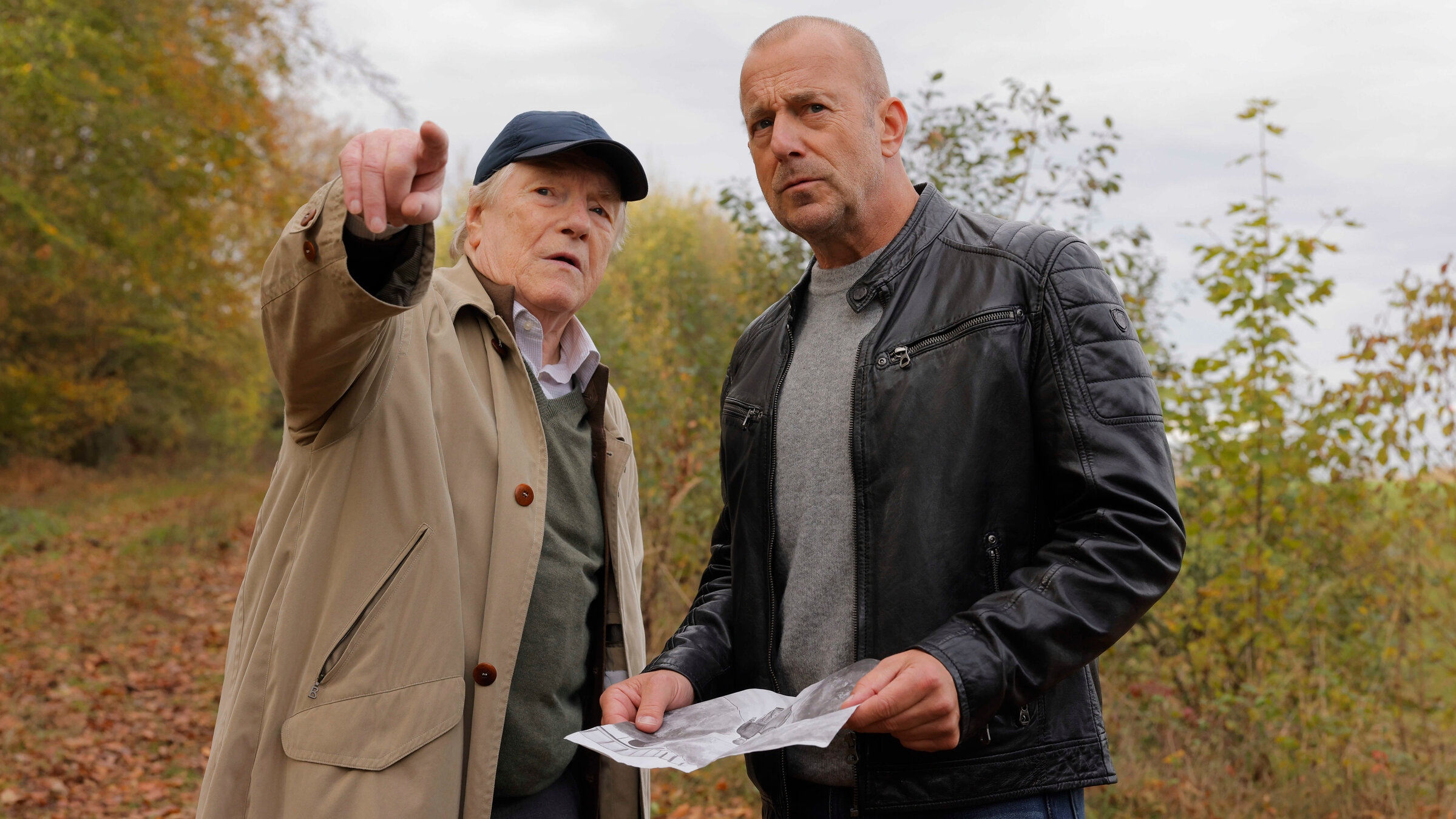 "Briefe aus dem Jenseits?": Gerd Dennert (Manfred Zapatka) und Ingo Thiel (Heino Ferch) stehen in der Natur. Ingo hat ein Bild in der Hand und und schaut aufmerksam in die Richtung, in die Gerd zeigt, während er ihm etwas zu erklären scheint.