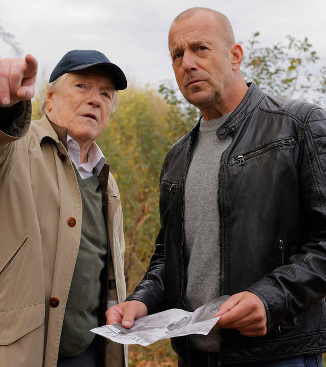"Briefe aus dem Jenseits?": Gerd Dennert (Manfred Zapatka) und Ingo Thiel (Heino Ferch) stehen in der Natur. Ingo hat ein Bild in der Hand und und schaut aufmerksam in die Richtung, in die Gerd zeigt, während er ihm etwas zu erklären scheint.