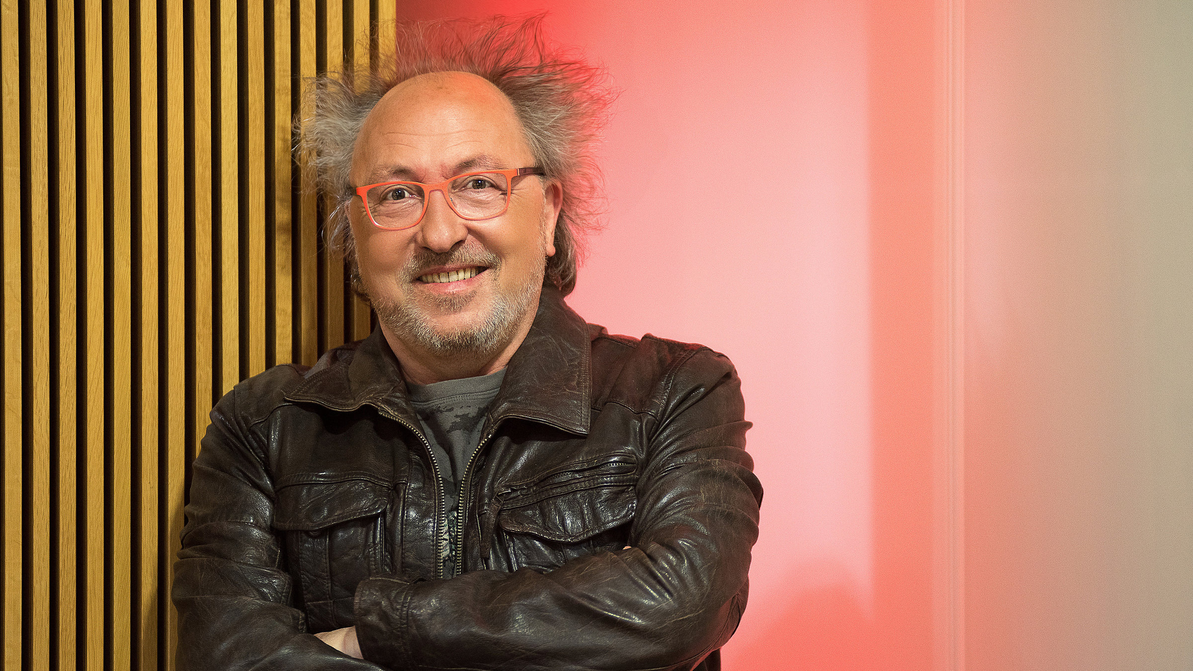 "Urban Priol: Im Fluss": Urban Priol trägt eine schwarze Lederjacke und steht mit verschränkten Armen vor einer Holzwand. Er lächelt freundlich in die Kamera.