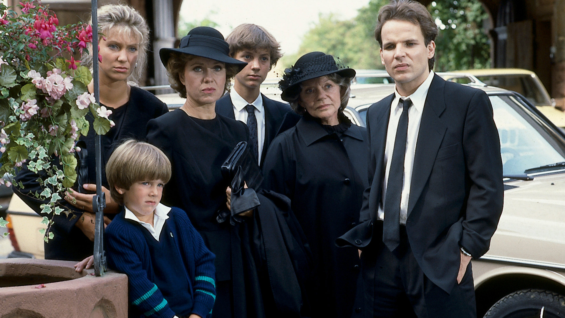 "Diese Drombuschs - In der Mitte des Lebens": Marion (Sabine Kaack), Vera Drombusch (Witta Pohl), Thomi (Eike Hagen Schweikhardt), Oma Magarete (Grete Wurm), Chris (Mick Werup) und Daniel (Jan Harndorf) stehen in Trauerkleidung auf einem Parkplatz und schauen betrübt.