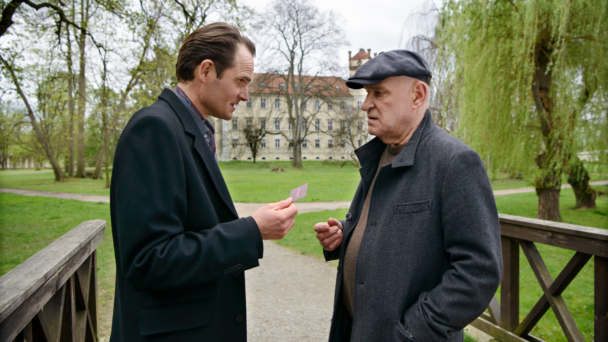 "Spreewaldkrimi - Böses muss mit Bösem enden": Kriminalrat a.D. Krüger (Christian Redl) und Prof. Levi A. Than (Fabian Hinrichs) unterhalten sich in einem Park. Prof. Than überreicht Krüger seine Visitenkarte.