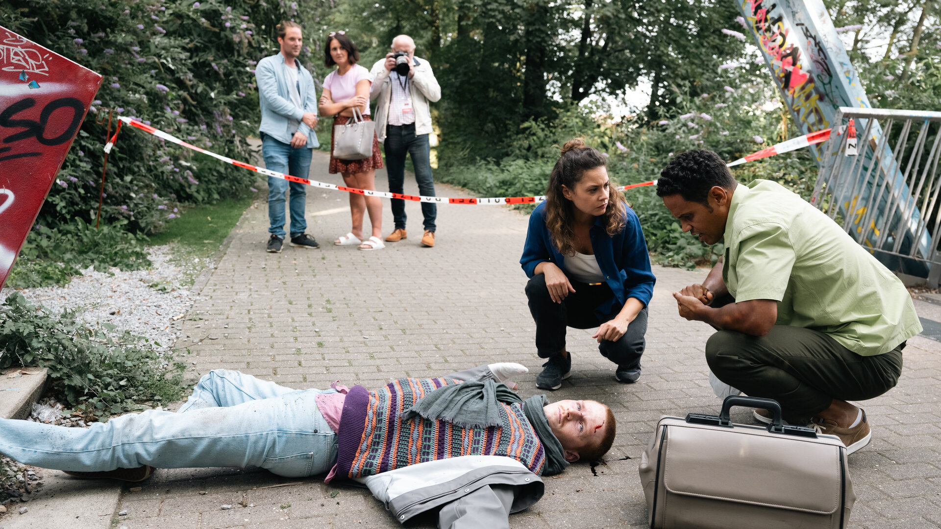 "SOKO Köln - Die letzte Aktion": Lilly Funke (Tatjana Kästel) kniet neben Dr. Adam Makena (Stephen Appleton) an einem Tatort auf Asphaltboden. Vor ihnen liegt der tote Marten Keller (Cedric Sprick-Benz) auf dem Rücken. Seine Arme sind zur Seite gespreizt und an seinem Kopf befindet sich Blut. Lilly sieht Adam ernst an. Dieser blickt nachdenklich auf den Toten. Im Hintergrund stehen hinter Polizeiabsperrband auf dem Gehweg zwei Männer (Komparsen) und eine Frau (Komparsin). Einer der neugierigen Passanten fotografiert mit einer Kamera den Tatort.