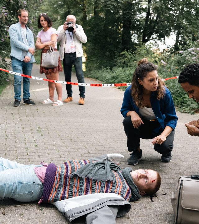 "SOKO Köln - Die letzte Aktion": Lilly Funke (Tatjana Kästel) kniet neben Dr. Adam Makena (Stephen Appleton) an einem Tatort auf Asphaltboden. Vor ihnen liegt der tote Marten Keller (Cedric Sprick-Benz) auf dem Rücken. Seine Arme sind zur Seite gespreizt und an seinem Kopf befindet sich Blut. Lilly sieht Adam ernst an. Dieser blickt nachdenklich auf den Toten. Im Hintergrund stehen hinter Polizeiabsperrband auf dem Gehweg zwei Männer (Komparsen) und eine Frau (Komparsin). Einer der neugierigen Passanten fotografiert mit einer Kamera den Tatort.