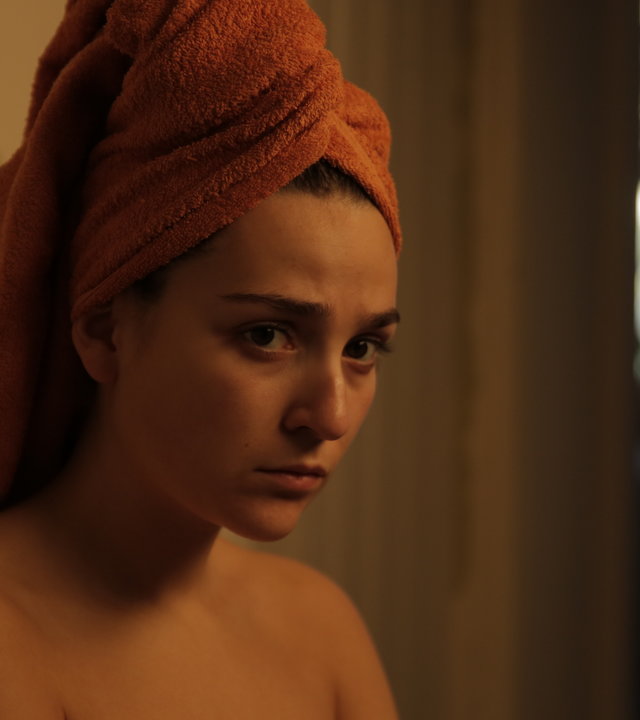 "About Saturday - Screw you guys I'm going home": Klara (Darin Hagi) steht mit einem Handtuch bekleidet in einem Flur und beobachtet etwas.