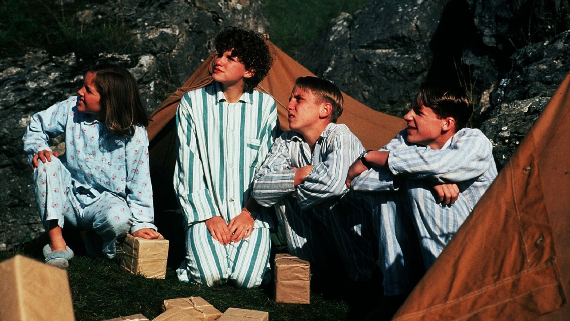 "Fünf Freunde im Nebel": Anne (Laura Petela), George (Jemima Rooper), Dick (Paul Child) und Julian (Marco Williamson) sitzen in Schlafanzügen zwischen zwei Zelten. Diese sind auf einem Rasen aufgebaut. Im Hintergrund sind Felsen zu erkennen.