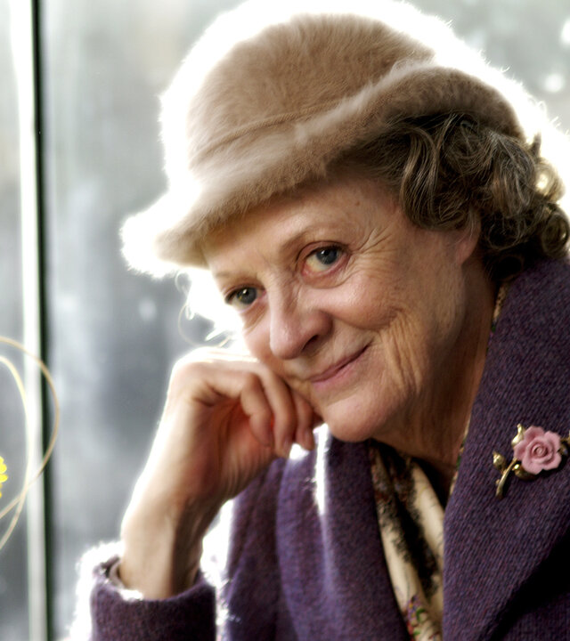 "Mord im Pfarrhaus": Grace (Maggie Smith) sitzt lächelnd an einem Fenster und stützt ihren Kopf auf ihre Hand. Sie trägt einen beigefarbenen Hut und einen violetten Mantel mit Blumenbrosche. Neben ihr steht eine Vase mit gelben Blumen.