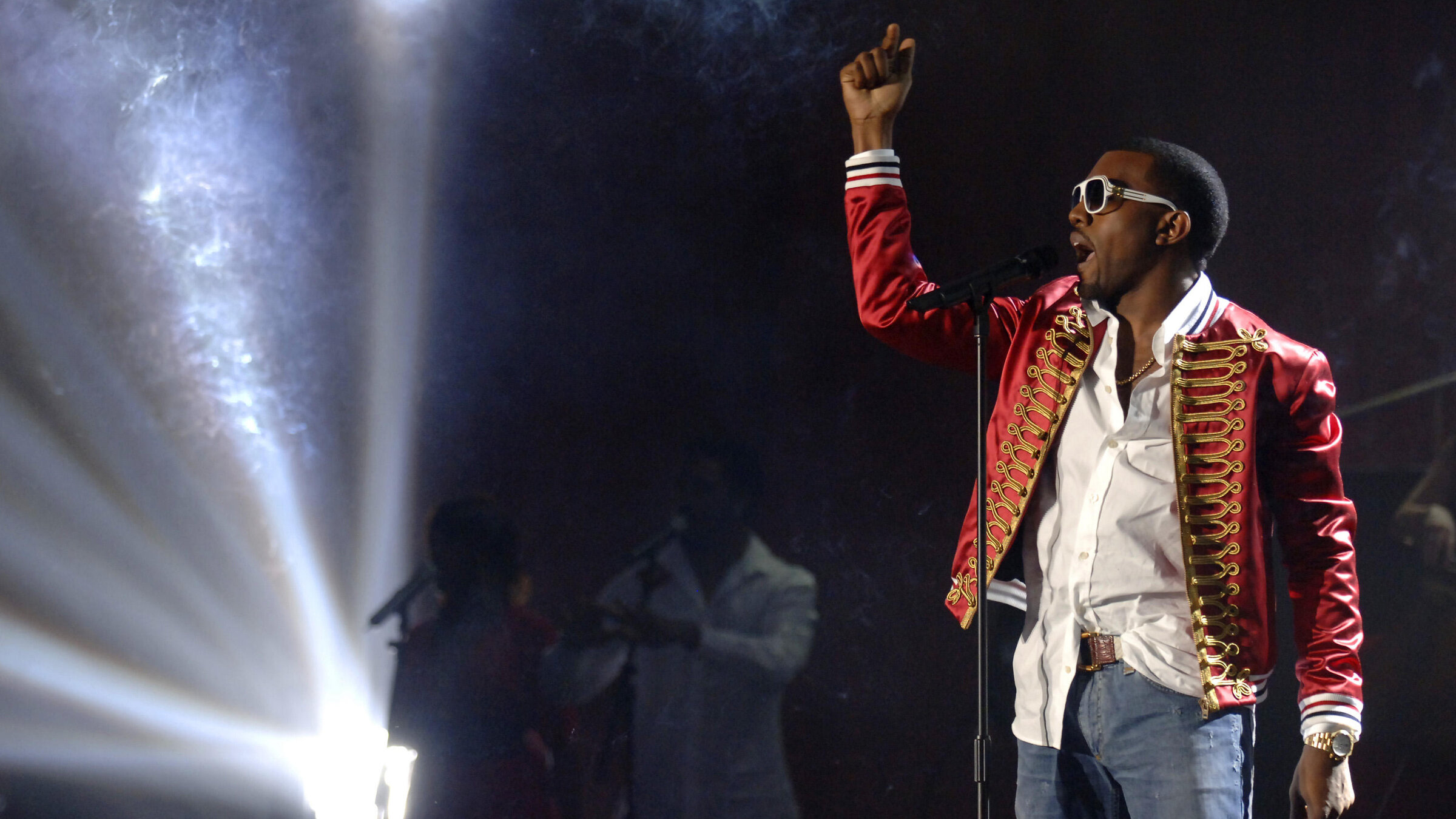 "The True Story of Kanye West": Bühne mit grellem Scheinwerferlicht links. Rechts ein Mann in Jeans, weißem Hemd und rotem Zirkus-Sakko, trägt Sonnenbrille