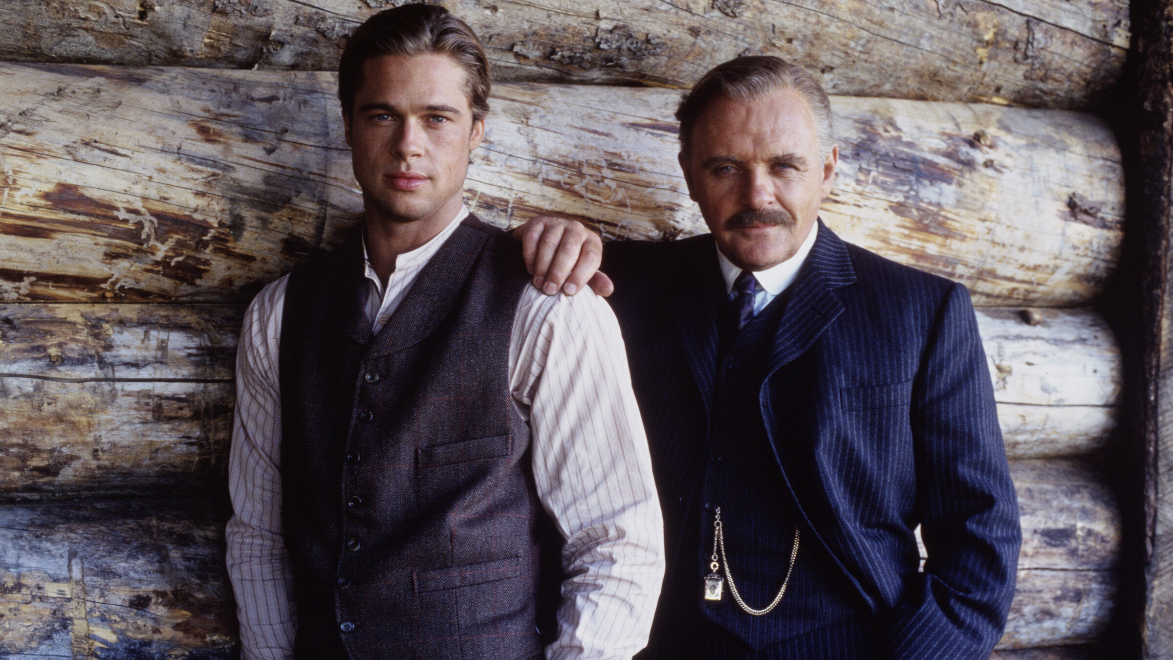 "Legenden der Leidenschaft": Tristan Ludlow (Brad Pitt) und Colonel William Ludlow (Anthony Hopkins) tragen Anzüge mit Weste und posieren lässig in die Kamera blickend nebeneinander vor einer Blockhauswand. Tristan ist ohne Sakko, sein Vater hat eine Hand auf seine Schulter gelegt.