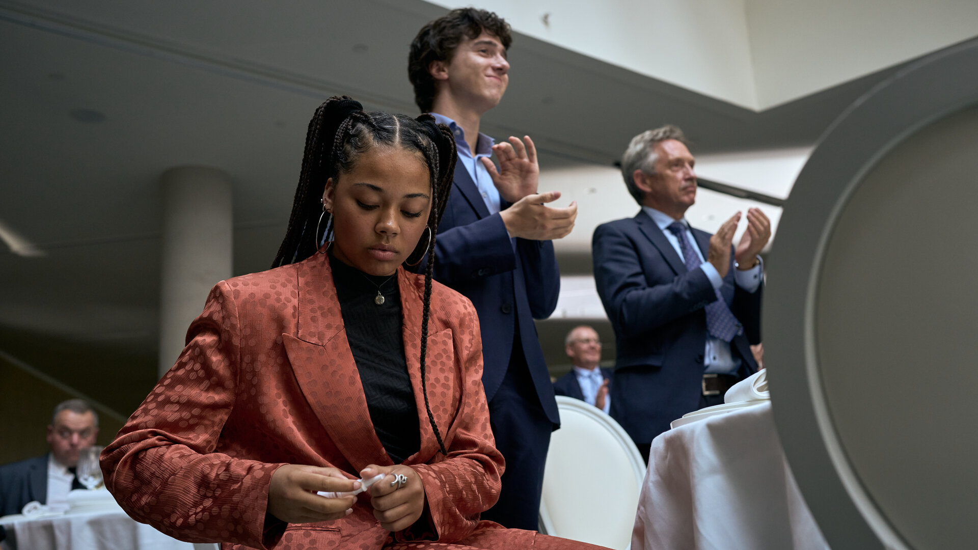 "Elixir – The Pain Killers - Die Islandmuschel": Lola (J-Leeta Tijmes) sitzt in einem Raum an einem Tisch mit weißer Tischdecke. Sie schaut auf ihre Hände. Art (Roman Derwig) und ein weiterer Mann (Komparse) stehen neben ihr und applaudieren. Im Hintergrund befinden sich zwei weitere Personen (Komparsen).