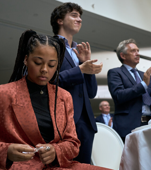 "Elixir – The Pain Killers - Die Islandmuschel": Lola (J-Leeta Tijmes) sitzt in einem Raum an einem Tisch mit weißer Tischdecke. Sie schaut auf ihre Hände. Art (Roman Derwig) und ein weiterer Mann (Komparse) stehen neben ihr und applaudieren. Im Hintergrund befinden sich zwei weitere Personen (Komparsen).