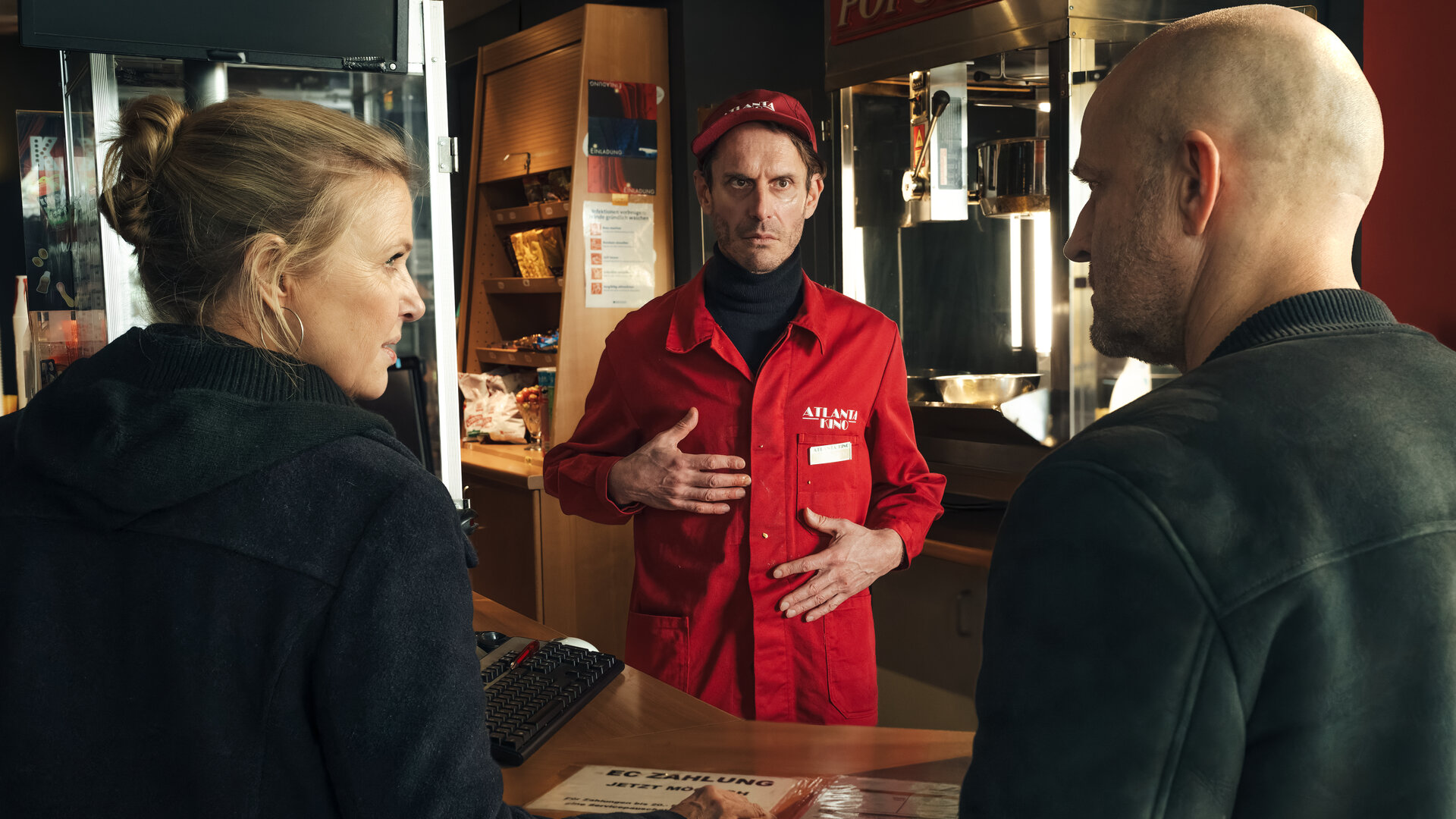"Die Chefin - Dunkelfeld (2)": Vor einer Kino-Theke stehen Vera Lanz (Katharina Böhm) und Robert Heffler (Jürgen Vogel). Beide sehen sich an. Hinter der Theke, in roter Arbeitskleidung, steht Thorsten Handke (Max Mayer), der beide Hände vor seinen Oberkörper hält.