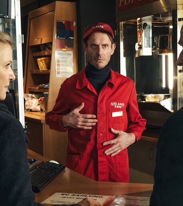 "Die Chefin - Dunkelfeld (2)": Vor einer Kino-Theke stehen Vera Lanz (Katharina Böhm) und Robert Heffler (Jürgen Vogel). Beide sehen sich an. Hinter der Theke, in roter Arbeitskleidung, steht Thorsten Handke (Max Mayer), der beide Hände vor seinen Oberkörper hält.