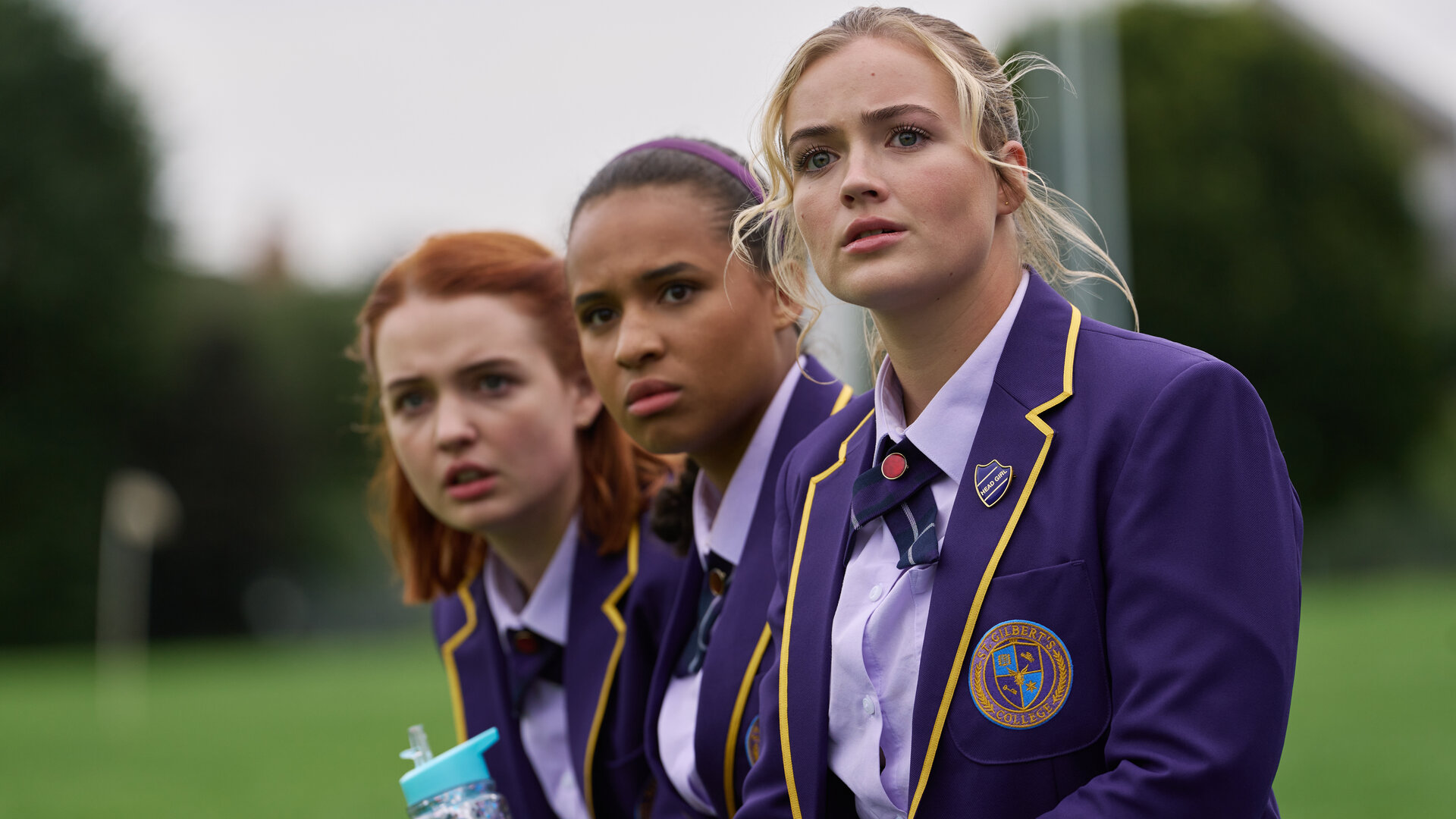 "Boarders - Welcome to St. Gilbert's - Party & Payback": Beatrix (Tallulah Greive), Abby (Assa Kanouté) und Florence (Rosie Graham) sitzen nebeneinander und beobachten etwas. Sie tragen ihre Schuluniformen. Im Hintergrund ist eine Rasenfläche zu erkennen.