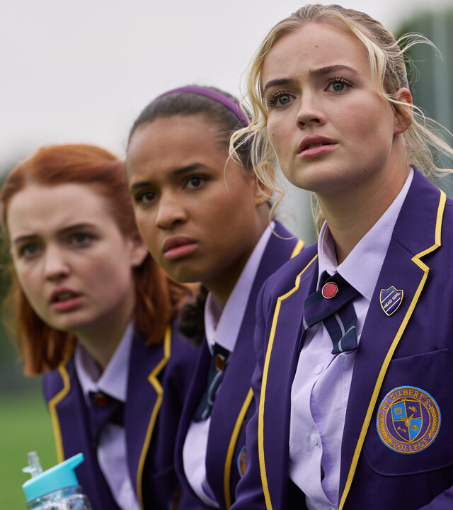 "Boarders - Welcome to St. Gilbert's - Party & Payback": Beatrix (Tallulah Greive), Abby (Assa Kanouté) und Florence (Rosie Graham) sitzen nebeneinander und beobachten etwas. Sie tragen ihre Schuluniformen. Im Hintergrund ist eine Rasenfläche zu erkennen.