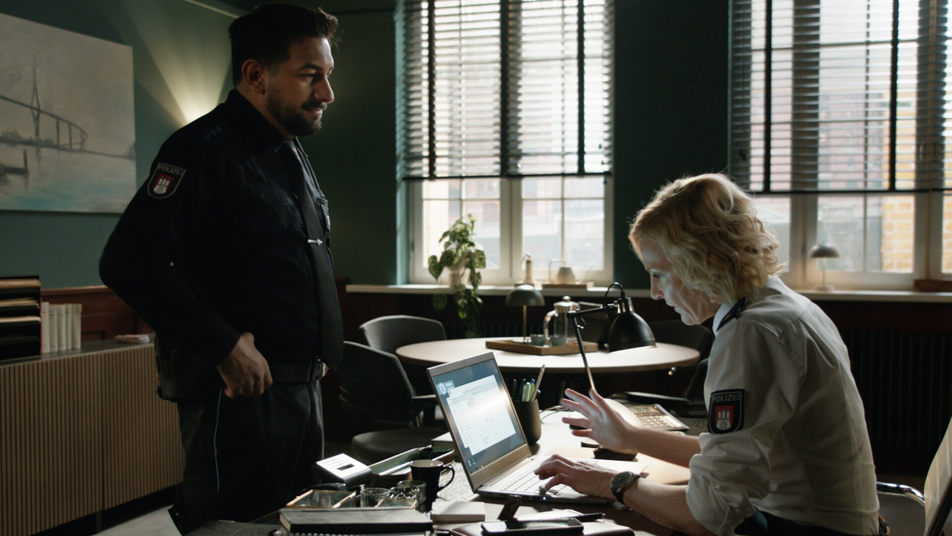 "Notruf Hafenkante - Vertrauensbruch": Die Polizistin Melanie Hansen (Sanna Englund) sitzt in ihrem Büro an ihrem Schreibtisch. Sie tippt etwas in einen Laptop, der geöffnet vor ihr steht. Vor ihrem Schreibtisch steht ihr Kollege Malik (Zejhun Demirov). Er grinst sie an.